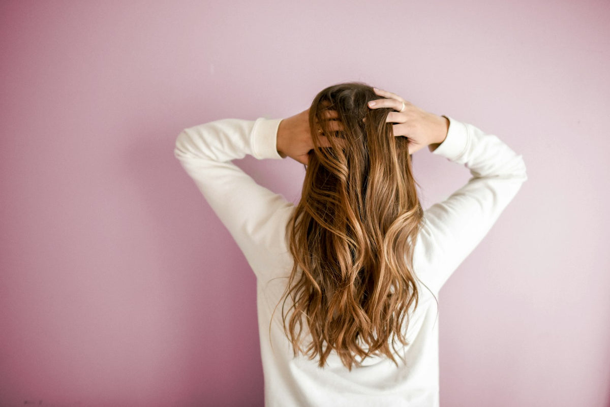 A woman with long, soft, conditioned wavy hair seen from behind against a pink wall
