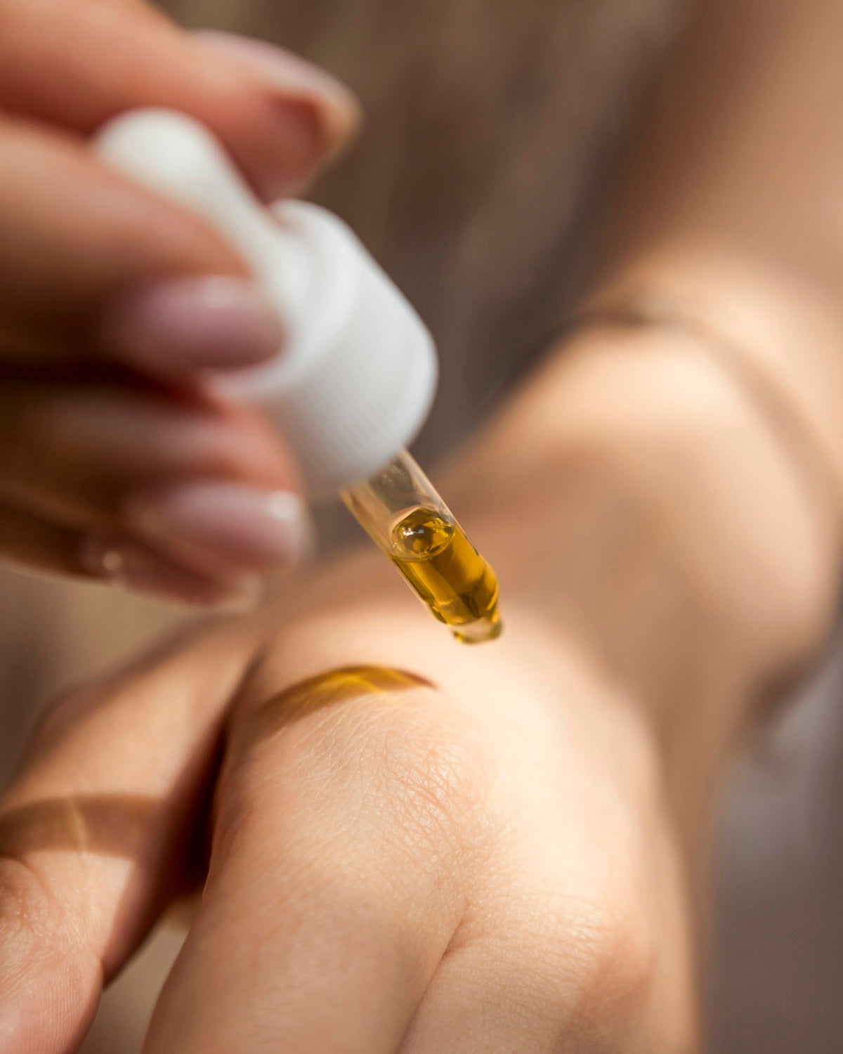 A hand releasing a drop of golden serum from a glass dropper onto the back of another hand