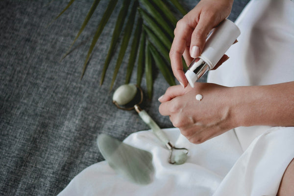 Hands applying lotion beside a green stone face roller and gua sha tool on white linen
