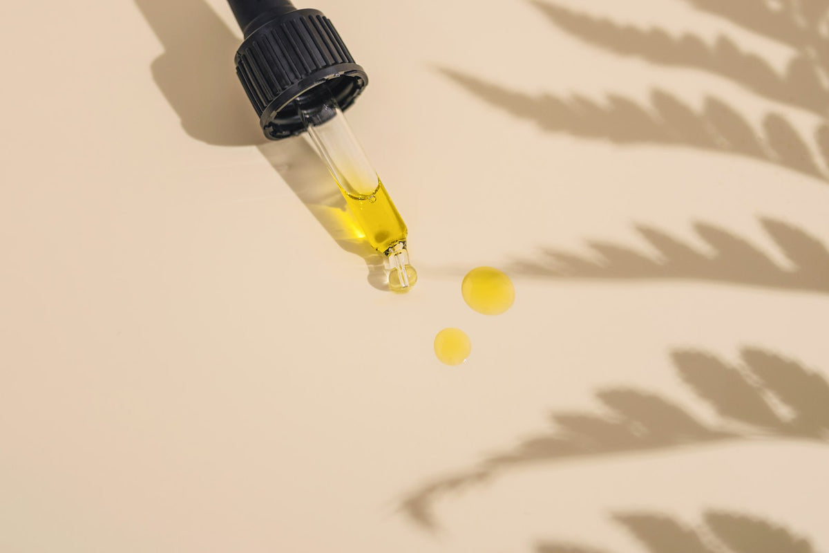 A glass dropper releasing drops of golden face oil for dry skin onto a beige surface with fern shadows
