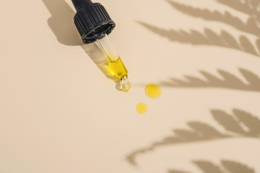 A glass dropper releasing drops of golden face oil for dry skin onto a beige surface with fern shadows
