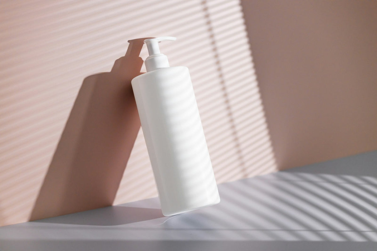A plain white pump bottle of sulfate-free shampoo standing against a pink wall in soft light