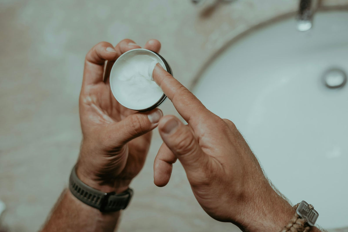 Hands scooping white magnesium balm from a small metal tin over a bathroom sink