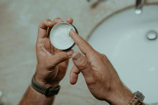 Hands scooping white magnesium balm from a small metal tin over a bathroom sink