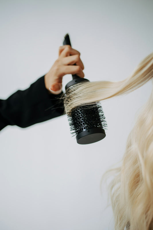 A stylist lifting long blonde hair with a round brush while blow-drying, a finishing step after volumizing shampoo